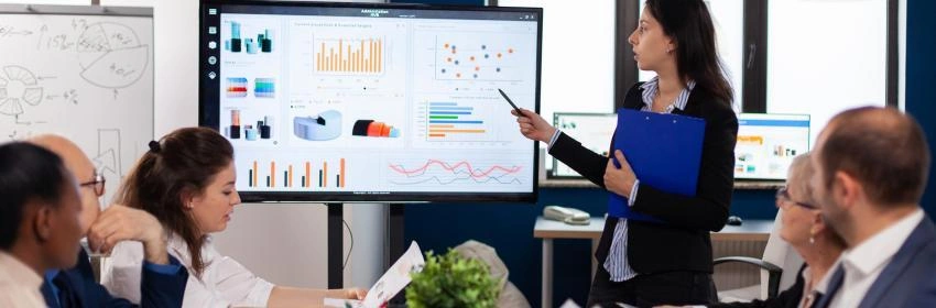 moça com terno preto fazendo apresentação com tela com planilhas, em um sala de reunião com várias pessoas